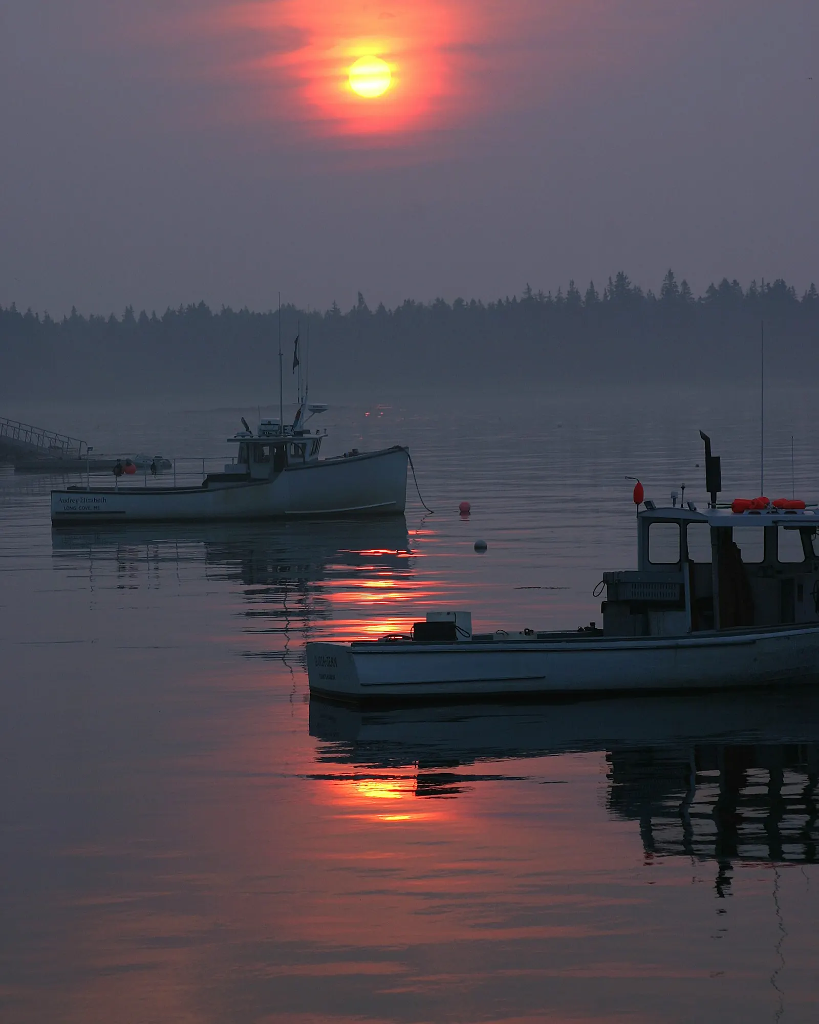 A small fishing boat moored on glassy water under a hazy red sunrise, the far shore a dark line of trees.
