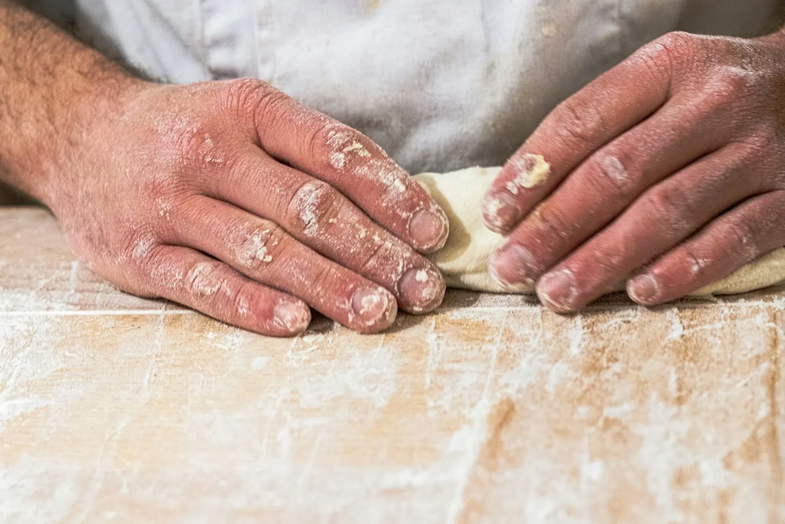 A baker's flour-dusted hands folding a pale slab of dough on a wooden counter.