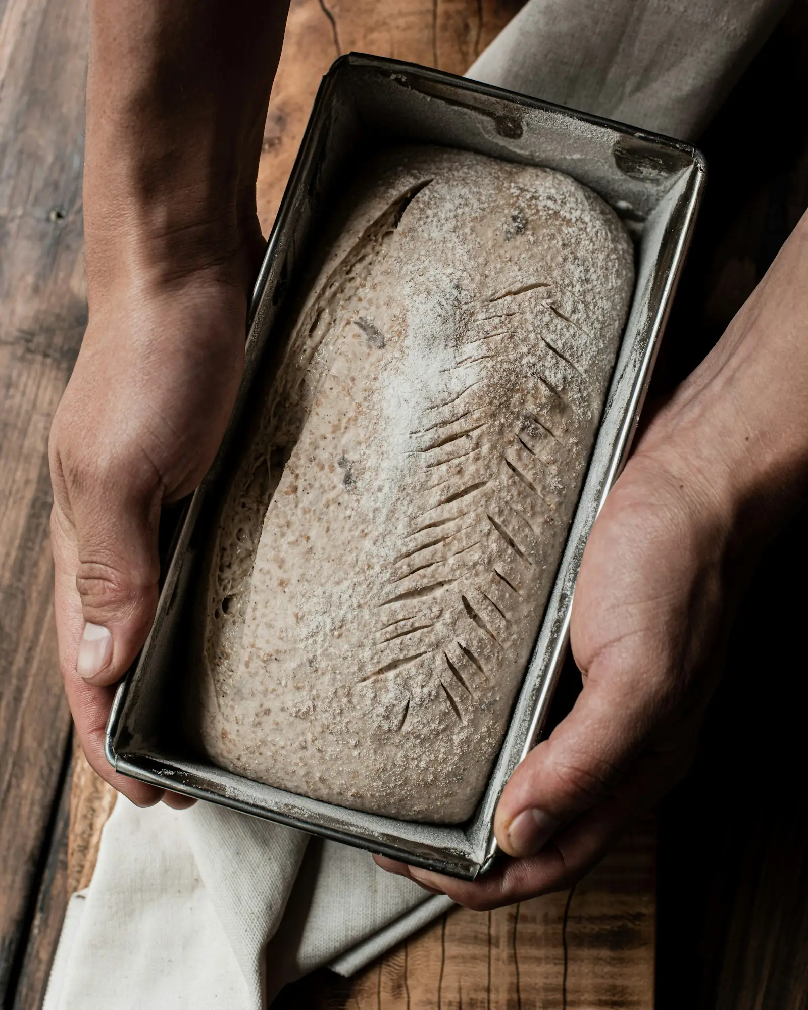 Two hands holding a dark rye loaf in a metal tin, the crust scored with a single long slash, on a scrubbed wooden board.