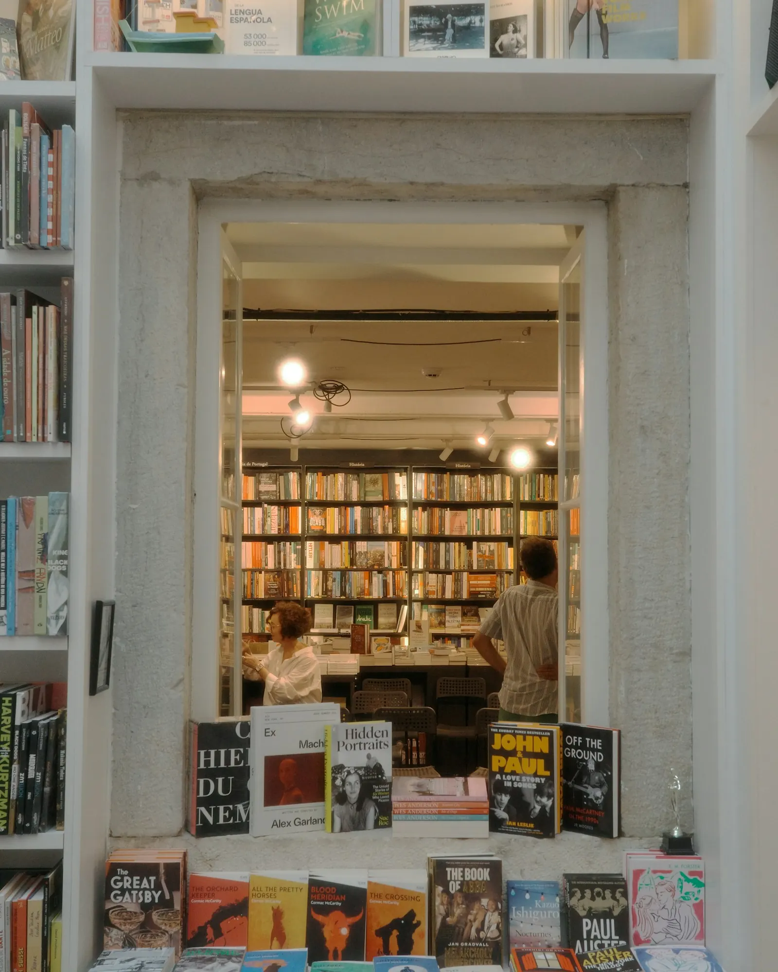 A narrow bookshop doorway framed by white shelves, new paperbacks fanned on a table in front and a second room of floor-to-ceiling books beyond.